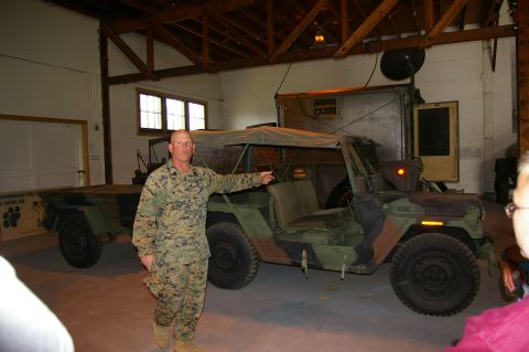 1986 Ford Bronco - A man in a military uniform stands next to a camouflaged military jeep and trailer inside a building.