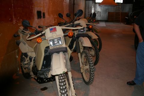 1986 Ford Bronco - Two tan military motorcycles, one with USMC markings, are parked indoors. The registry claims they are 1986 Ford Broncos, but this is incor…