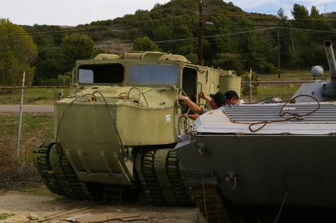 1986 Ford Bronco - Two individuals are interacting with the door of a green tracked amphibious vehicle parked next to another vehicle.