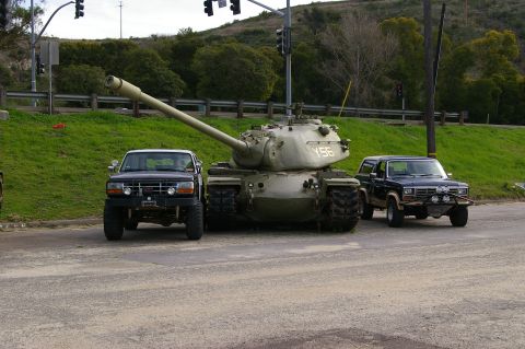 1986 Ford Bronco - A military tank is parked between two SUVs on a paved road with a grassy hill in the background.