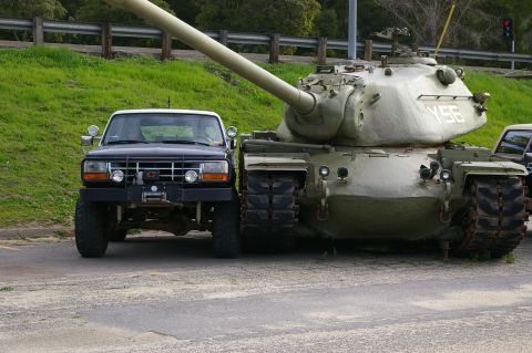 1986 Ford Bronco - A Ford truck is parked next to a military tank, with a second vehicle partially visible on the right.