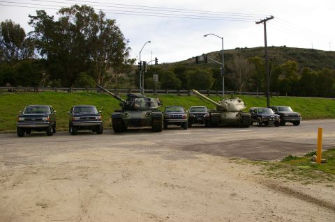 1986 Ford Bronco - A line of SUVs and two military tanks are parked in a lot with a grassy hill in the background.