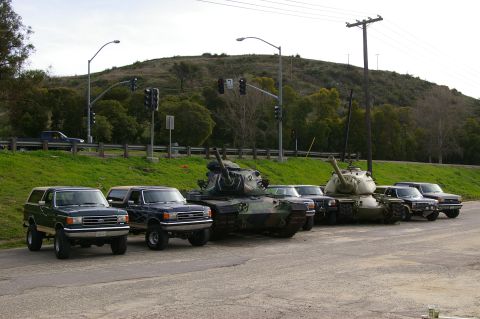 1986 Ford Bronco - Several Ford Broncos and military tanks are parked in a line on an asphalt lot.
