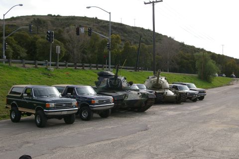 1986 Ford Bronco - A lineup of vehicles, including a Ford Bronco, pickup truck, and two tanks, are parked on the side of a road.