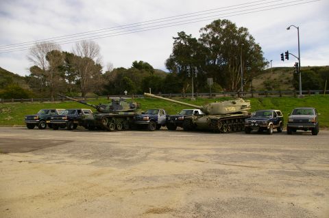 1986 Ford Bronco - Several military tanks and pickup trucks are parked in a line on a dirt lot under a cloudy sky.