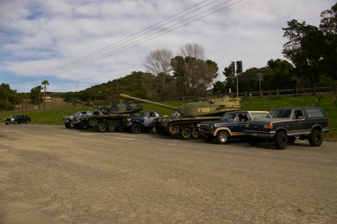 1986 Ford Bronco - A dark gray Ford Bronco is parked next to military tanks and pickup trucks.