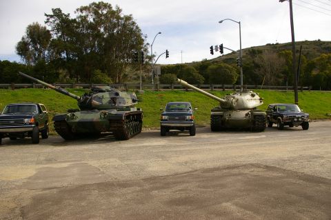 1986 Ford Bronco - Several military tanks and civilian trucks are parked side-by-side on a paved area with a grassy hill in the background.