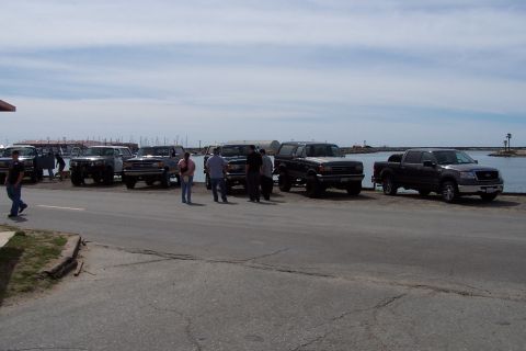 1990 Ford Bronco - A lineup of SUVs and trucks are parked along the waterfront, with boats visible in the harbor.