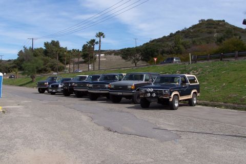1990 Ford Bronco - A line of Ford Broncos parked along a roadside with a grassy hill in the background.