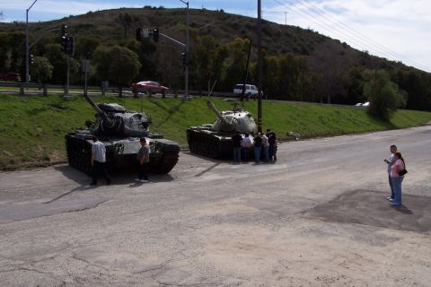 1990 Ford Bronco - Two military tanks are parked on a road next to a grassy embankment, with several people observing them.