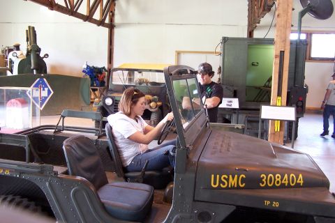 1990 Ford Bronco - A woman sits in the driver's seat of a USMC Jeep while a man stands behind her, looking into the vehicle.