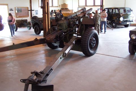 1990 Ford Bronco - Military vehicles and an artillery piece are displayed indoors at an event.