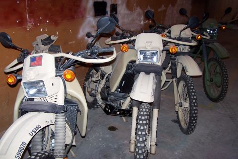 1990 Ford Bronco - Several military-style motorcycles are lined up in an indoor space, with two tan bikes in the foreground and a green one behind.