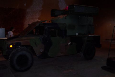 1990 Ford Bronco - A military-style vehicle with camouflage paint is parked indoors, with its hood open and equipment mounted on its roof.
