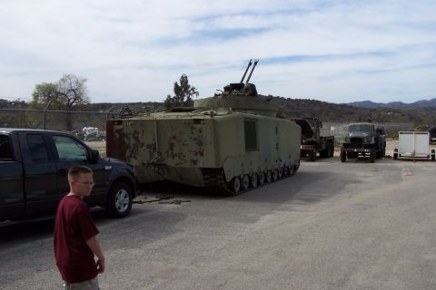 1990 Ford Bronco - An armored personnel carrier sits in a lot with other vehicles under a cloudy sky.