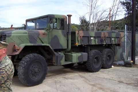 1990 Ford Bronco - A military truck with a camouflage paint scheme is parked on a dirt lot next to a chain-link fence.