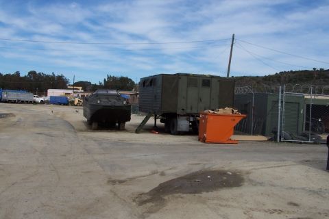 1990 Ford Bronco - Amphibious vehicle and trailer at an outdoor lot with a dumpster and other structures.