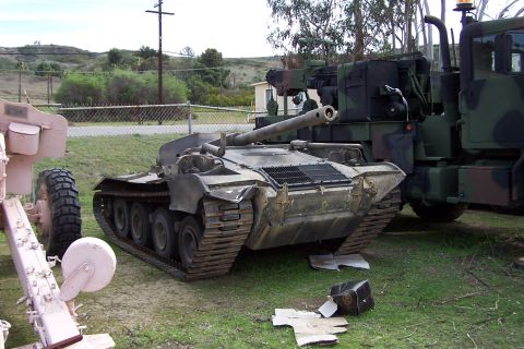 1990 Ford Bronco - A military tank sits on grass next to a camouflage truck, with debris scattered on the ground.
