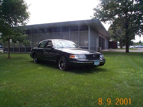 1998 Ford Crown Victoria - A black 1998 Ford Crown Victoria is parked on a grassy lawn in front of a modern building with large windows.