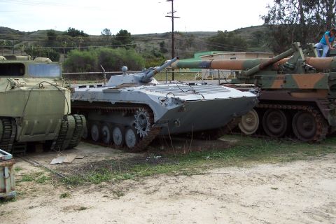 1990 Ford Bronco - Several military vehicles are parked in an outdoor lot, including a light gray tank and a camouflaged artillery piece.