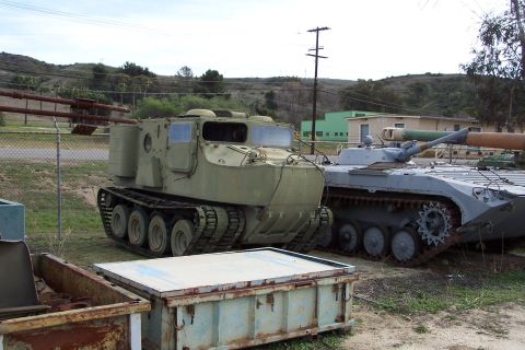 1990 Ford Bronco - Two tracked military vehicles, one green and one grey, are parked outdoors in a lot with buildings in the background.