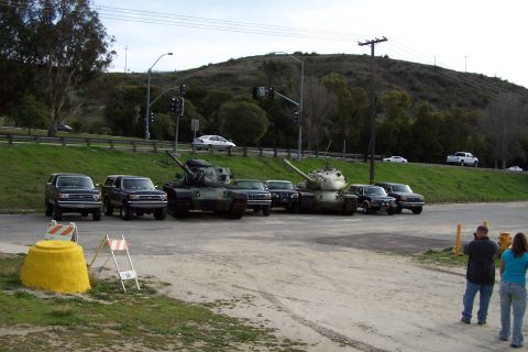 1990 Ford Bronco - Military tanks and SUVs are parked in a lot, with people observing in the foreground.