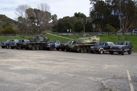 1990 Ford Bronco - Several military tanks and pickup trucks are parked in a lot, with trees and a hillside in the background.