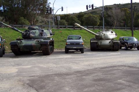 1990 Ford Bronco - A lineup of military tanks and civilian trucks are parked on a paved surface near a grassy embankment.