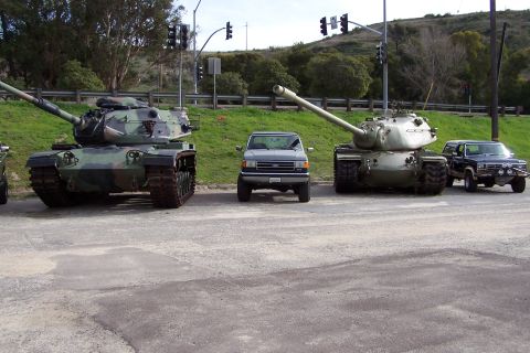 1990 Ford Bronco - A lineup of military tanks and SUVs are parked on a paved surface, with traffic signals visible overhead.