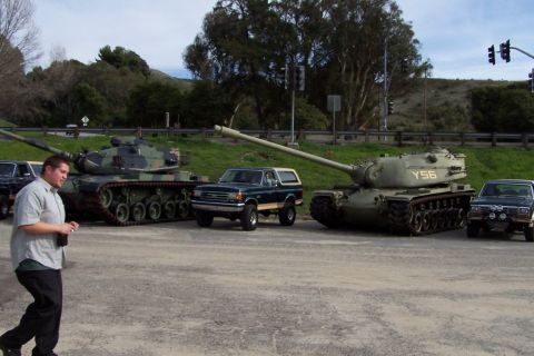 1990 Ford Bronco - A dark green 1990 Ford Bronco is parked between two military tanks at an outdoor event.
