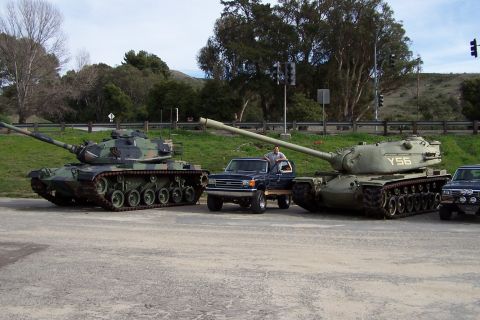 1990 Ford Bronco - Military tanks and a pickup truck parked outdoors with trees and a hillside in the background.