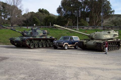 1990 Ford Bronco - A blue Ford Bronco is dwarfed by two large military tanks on a paved surface with grassy hills in the background.