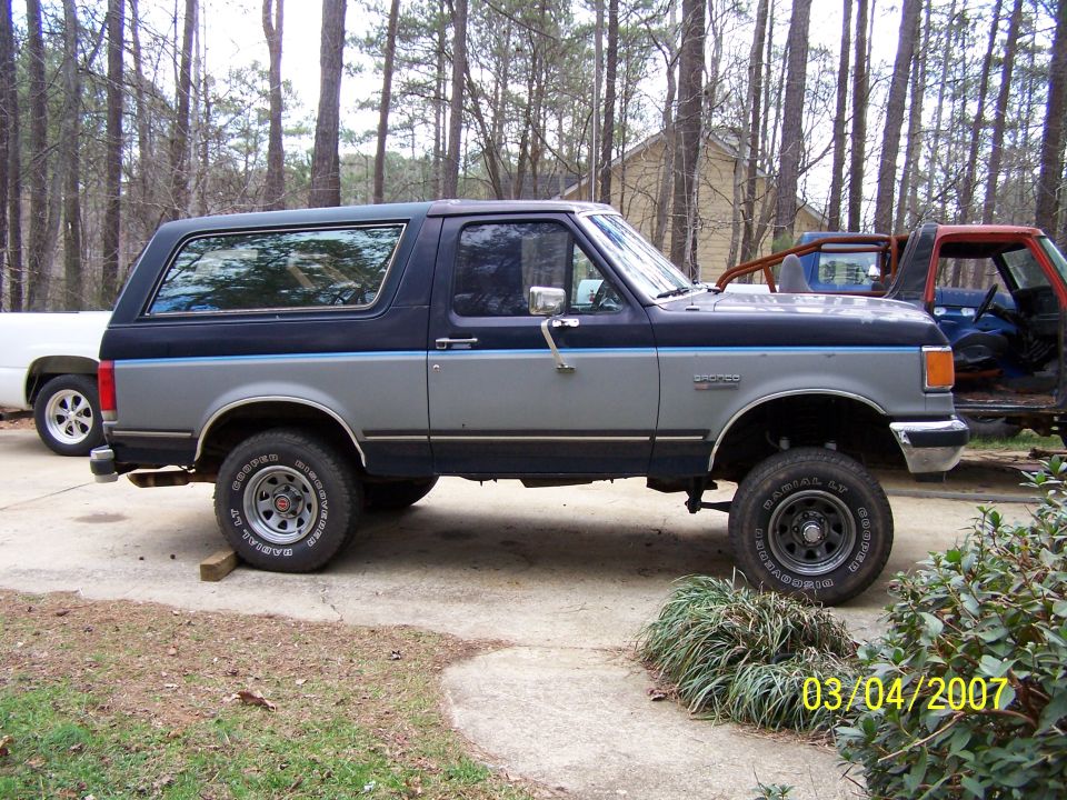 Side profile of a blue and silver Ford Bronco on Cooper Discoverer tires in a wooded driveway