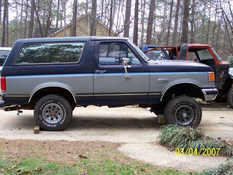 1979 Ford F-150 - Two Ford Broncos are parked outside on a concrete driveway. The front Bronco is dark blue and grey.