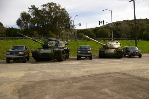 1986 Ford Bronco - Three SUVs are lined up in front of two military tanks on a sunny day.