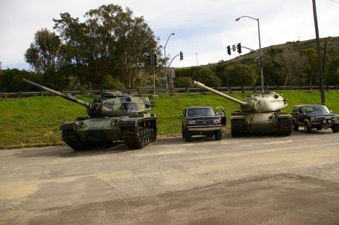 1986 Ford Bronco - A military convoy consisting of two tanks and three trucks is parked on the side of a road, with traffic lights visible in the background.