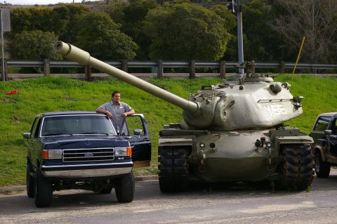 1986 Ford Bronco - A man stands by his Ford Bronco next to a large military tank.