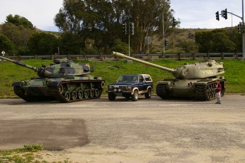 1986 Ford Bronco - A blue SUV is positioned between two military tanks on a paved surface with grass and trees in the background.