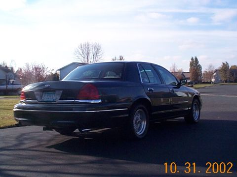 1998 Ford Crown Victoria - A dark 1998 Ford Crown Victoria sedan is viewed from the rear 3/4 angle on a paved surface.
