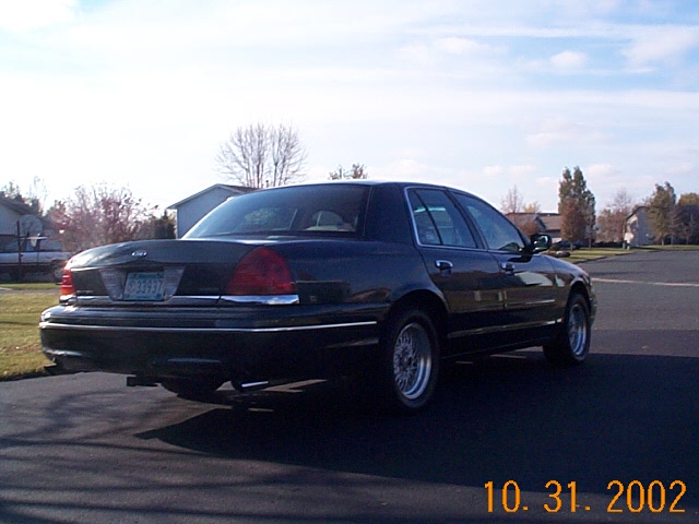 Dark 1998 Ford Crown Victoria sedan, rear 3/4 view, parked on asphalt with a grassy area to the left