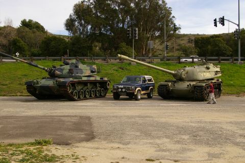 1986 Ford Bronco - A blue Ford Bronco is parked between two military tanks on a road.