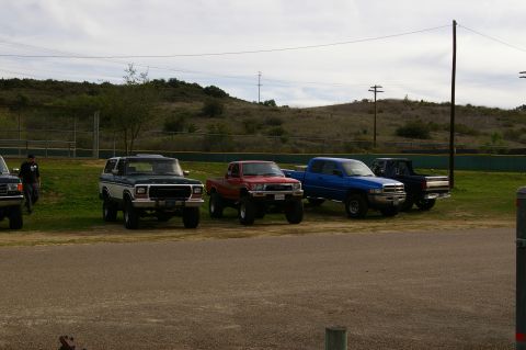 1986 Ford Bronco - Several trucks and an SUV are parked in a line on a grassy area next to a baseball field.