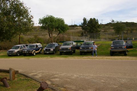 1986 Ford Bronco - A collection of SUVs and trucks are parked in a grassy area near a baseball field.
