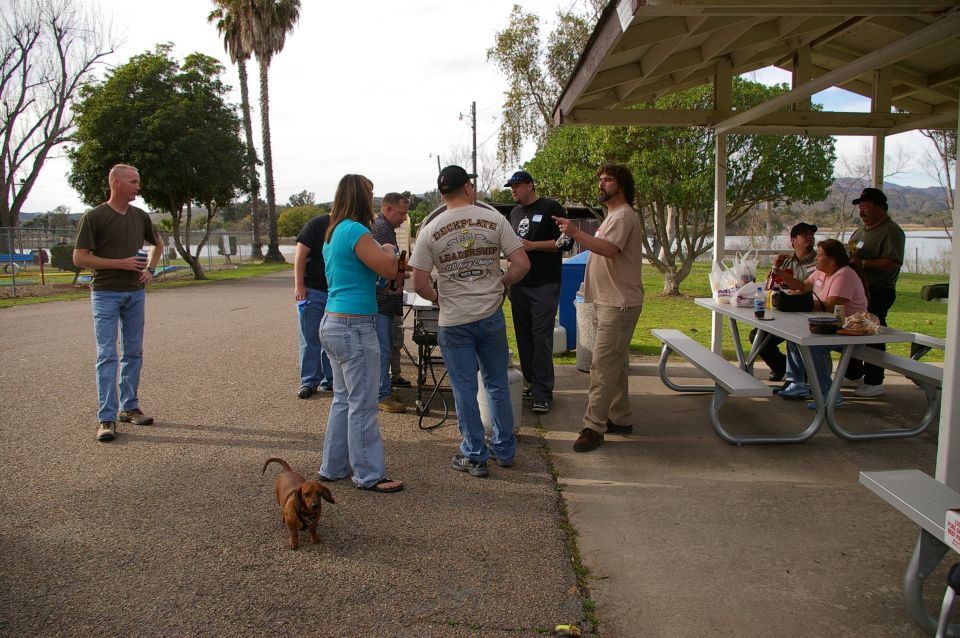 People gathered at picnic tables under a pavilion near a lake, with a dachshund in the foreground