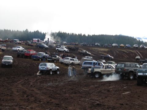 1996 Ford Bronco - A muddy field filled with off-road vehicles, with spectators gathered on a nearby hill.