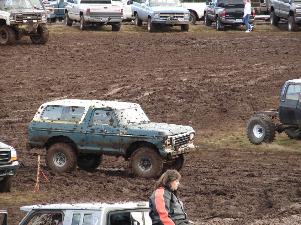 Mud-covered 1978 Ford Bronco with white top, stuck in mud pit. Other trucks visible in background