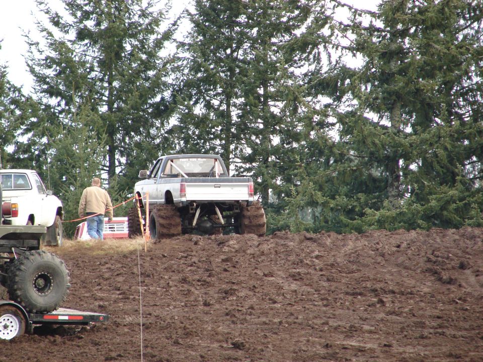 Pickup truck with huge tires in mud, another white truck nearby