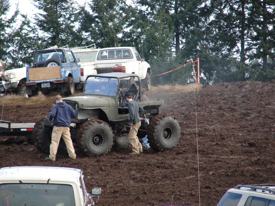 modified jeep with huge tires in mud, people around, other trucks in background