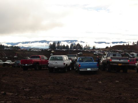 1996 Ford Bronco - A large gathering of trucks and SUVs are parked in a muddy field, with mountains visible in the distance under a cloudy sky.