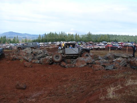 1996 Ford Bronco - Off-road buggy tackles a rocky obstacle course with spectators and parked vehicles in the background.